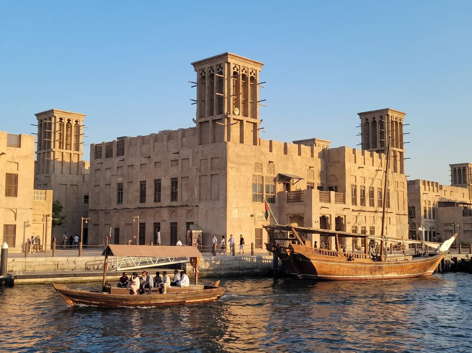 Al Seef'te rüzgâr kuleli kum rengi binaların önünde, Dubai Creek üzerinde yolcu teknesi ve büyük ahşap tekne görülüyor.