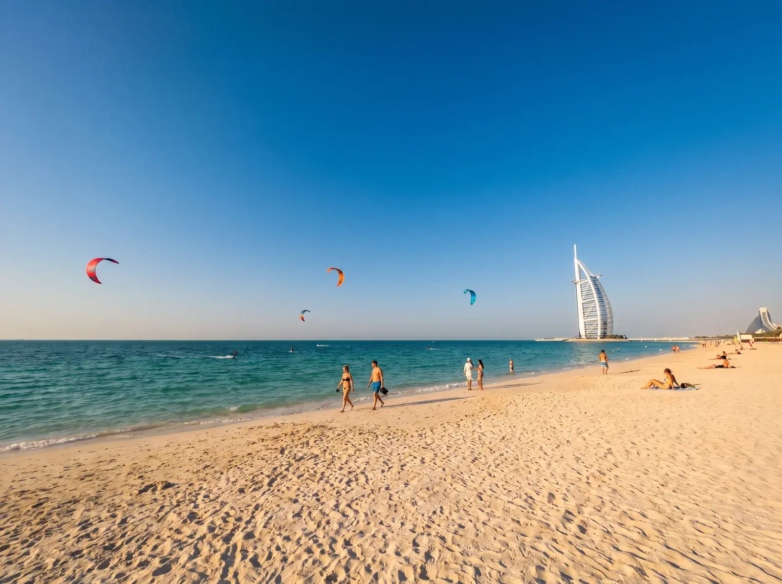 Kite Beach'te kumlu sahilde insanlar, denizin üstünde renkli uçurtmalar ve uzakta yelken biçimli Burj Al Arab görülüyor.