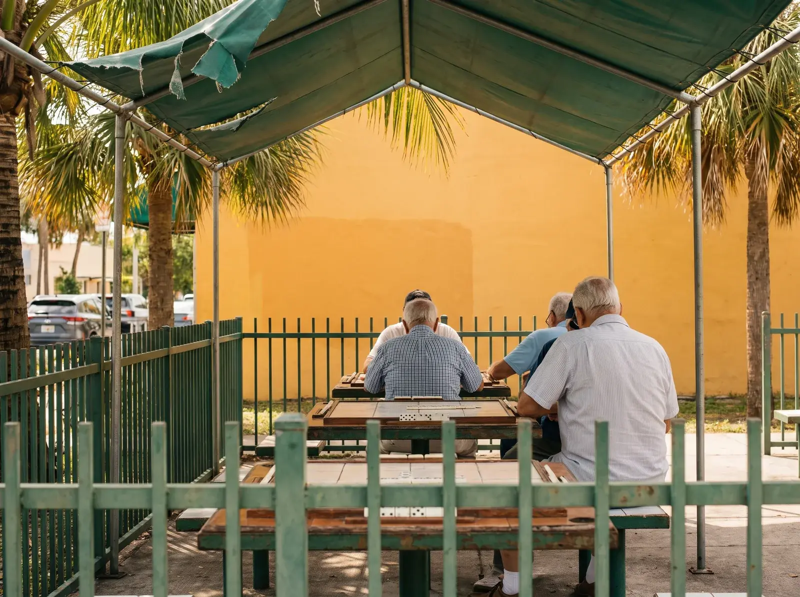 Little Havana'da yeşil tente altında masada oturan insanlar, yeşil parmaklıklar, palmiyeler ve sarı duvar görülüyor.