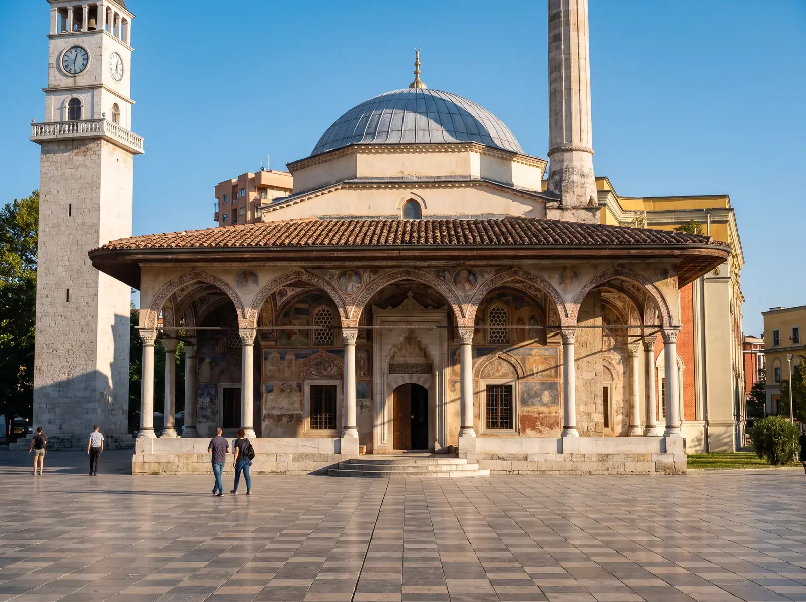 Tiran'da İskender Bey Meydanı'ndaki kemerli revaklı, kubbeli caminin yanında saat kulesi, önünde yürüyen insanlar görülüyor.