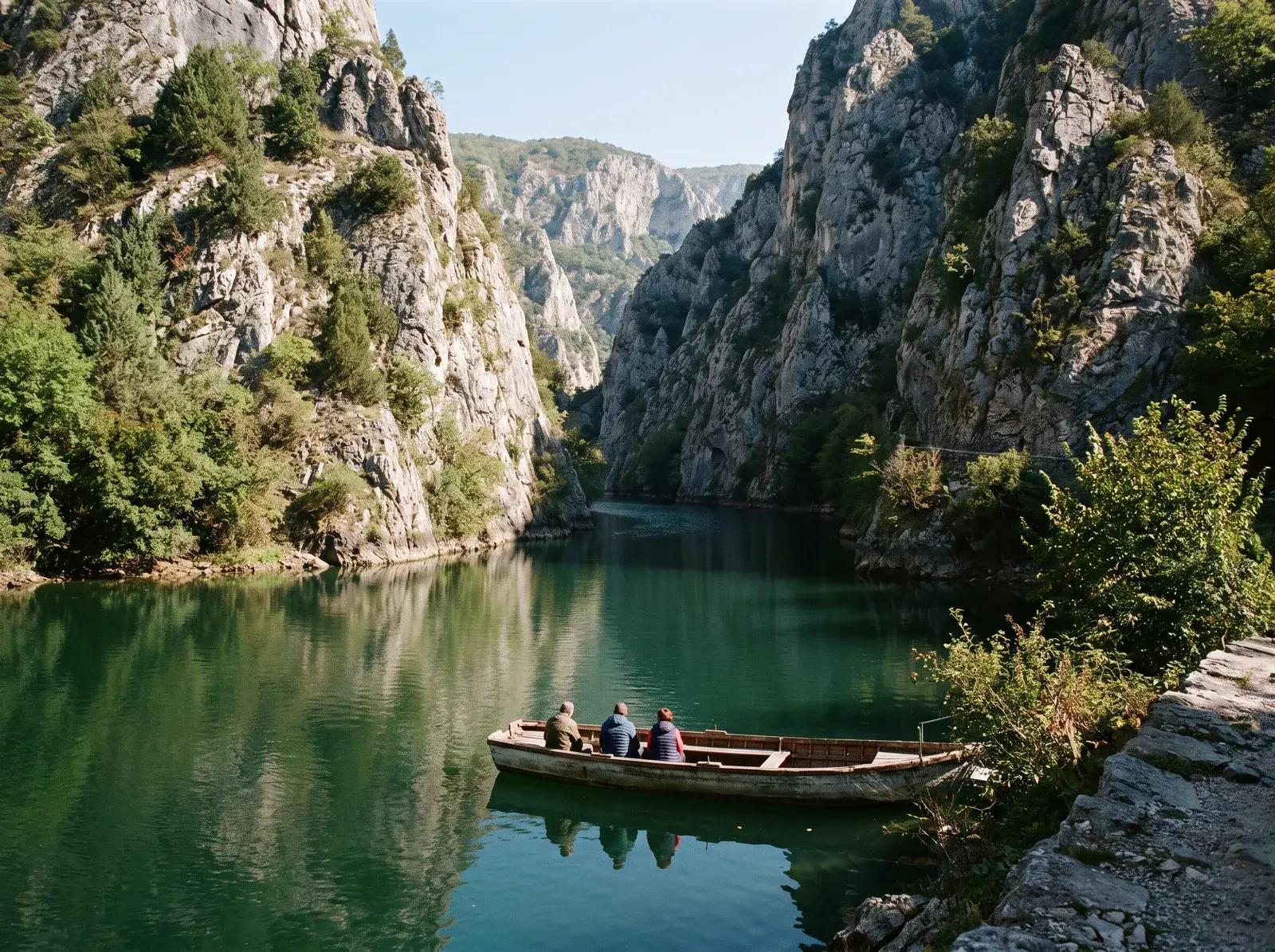 Matka Kanyonu'nda ağaçlarla kaplı dik kayalıklar yeşil suyu çevreliyor; kıyıya yakın bir kayıkta insanlar oturuyor.