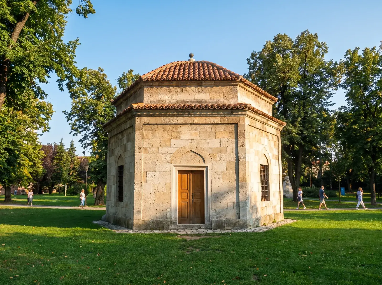 Damat Ali Paşa Türbesi'nin kiremit çatılı taş yapısı, çimenler, ağaçlar ve yürüyen insanlar arasında duruyor.