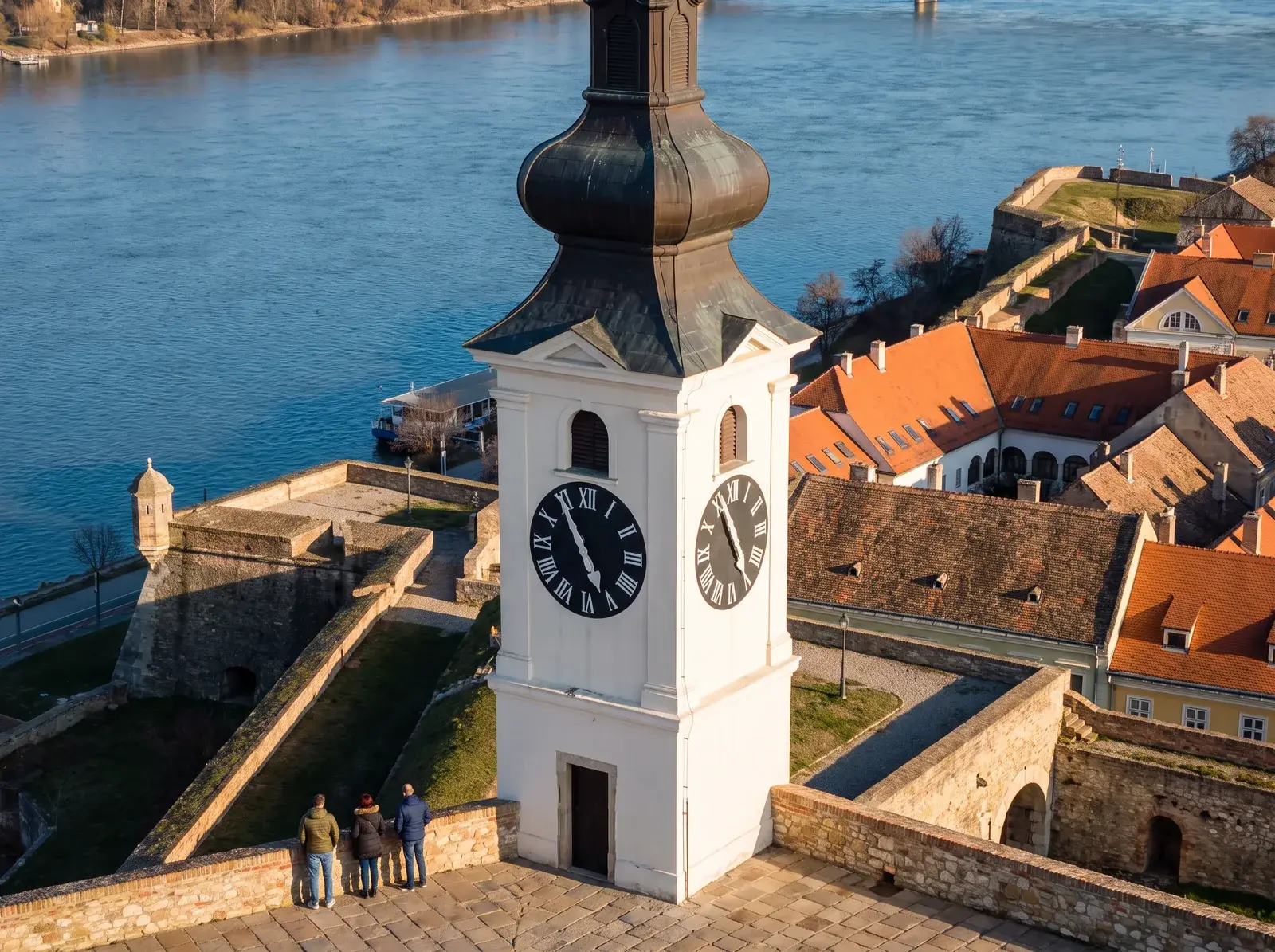 Petrovaradin Kalesi'nin beyaz saat kulesi, taş surlar ve kırmızı çatılar önünde, mavi nehrin kıyısında yükseliyor.