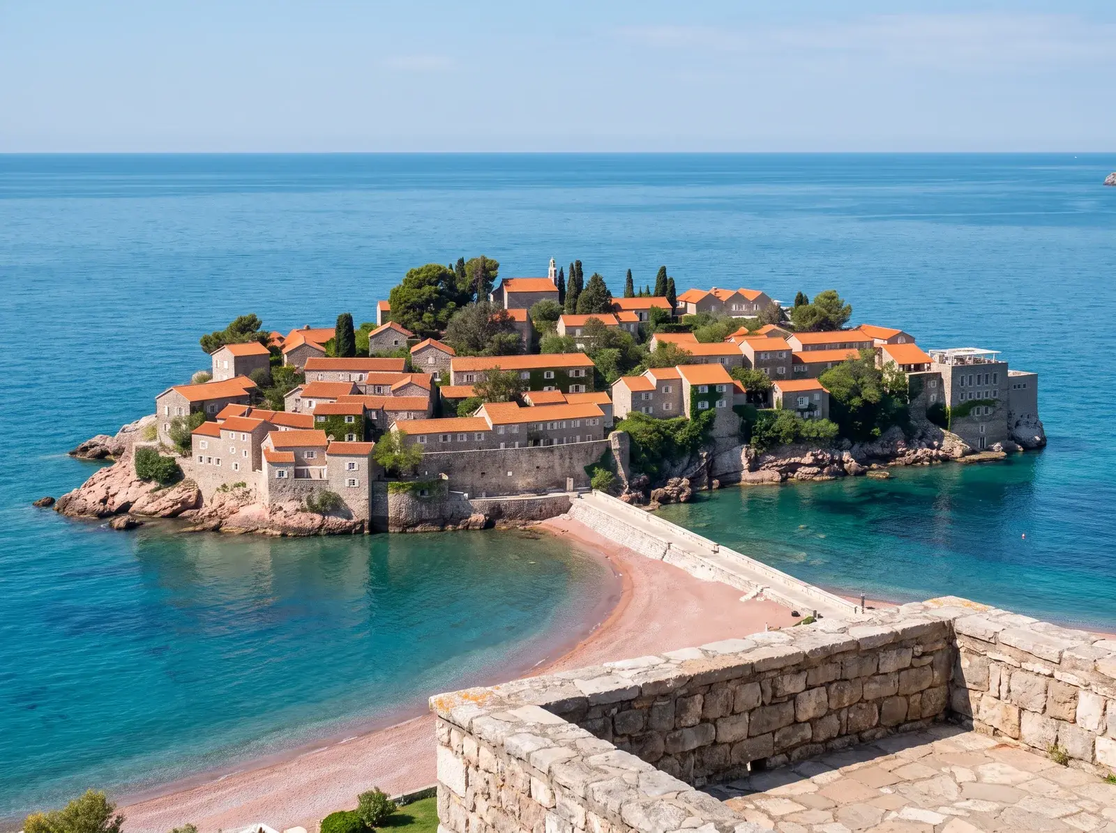 Sveti Stefan'ın kırmızı çatılı taş yapıları mavi denizle çevrili; dar bağlantı yolu kıyıdaki kumsala uzanıyor.