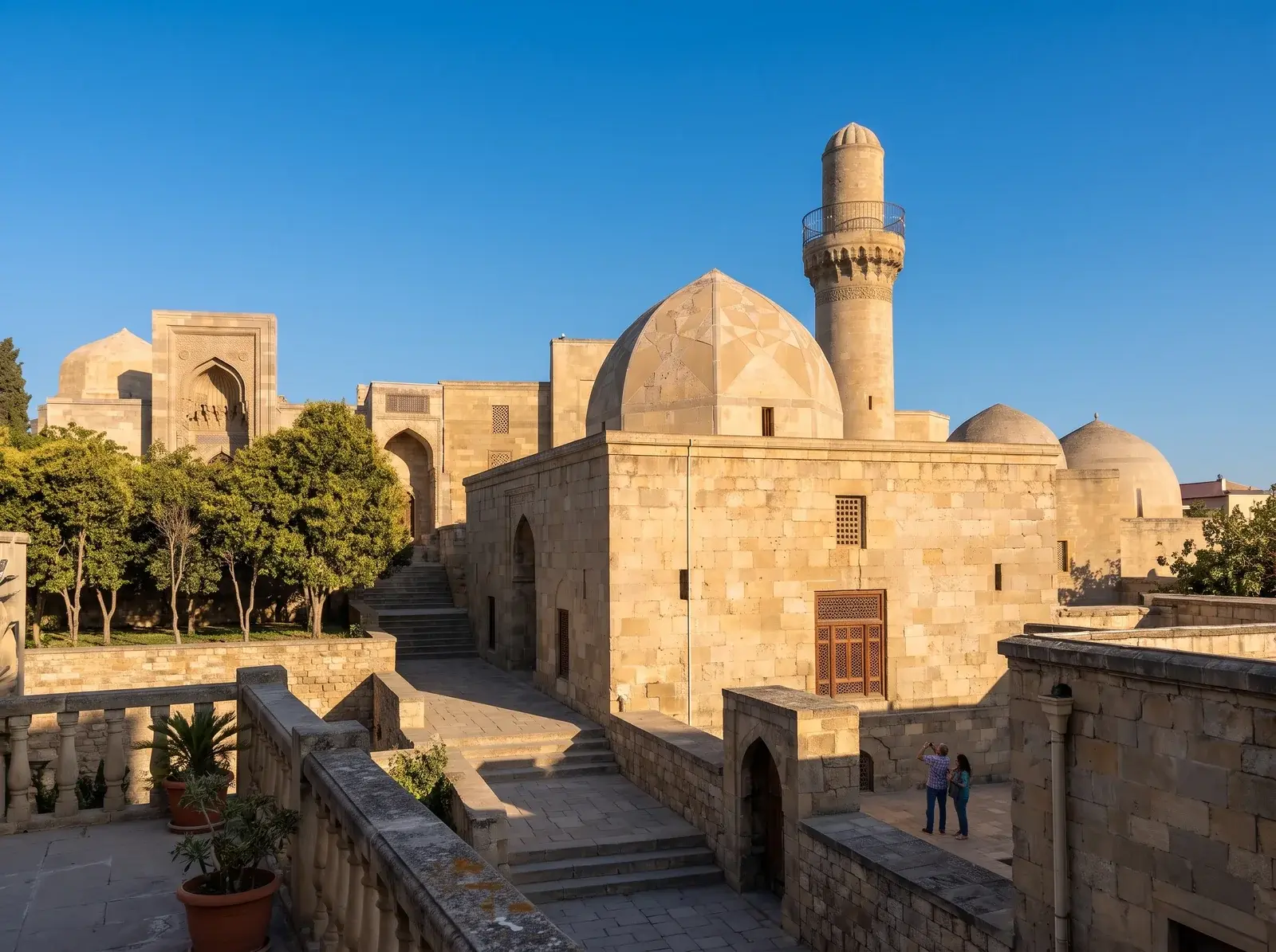 Şirvanşahlar Sarayı'nın taş yapıları, kubbesi ve minaresi, ağaçlı ve merdivenli avlunun üzerinde yükseliyor.
