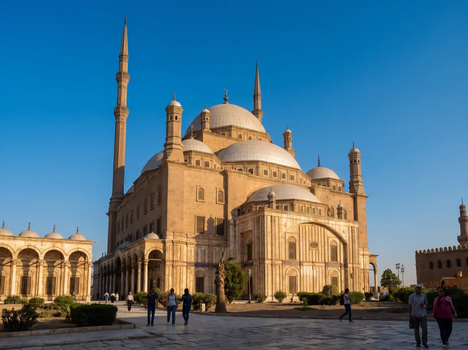 Muhammed Ali Camii'nin kubbeleri ve ince minareleri mavi gökyüzüne yükselirken taş avluda insanlar yürüyor.