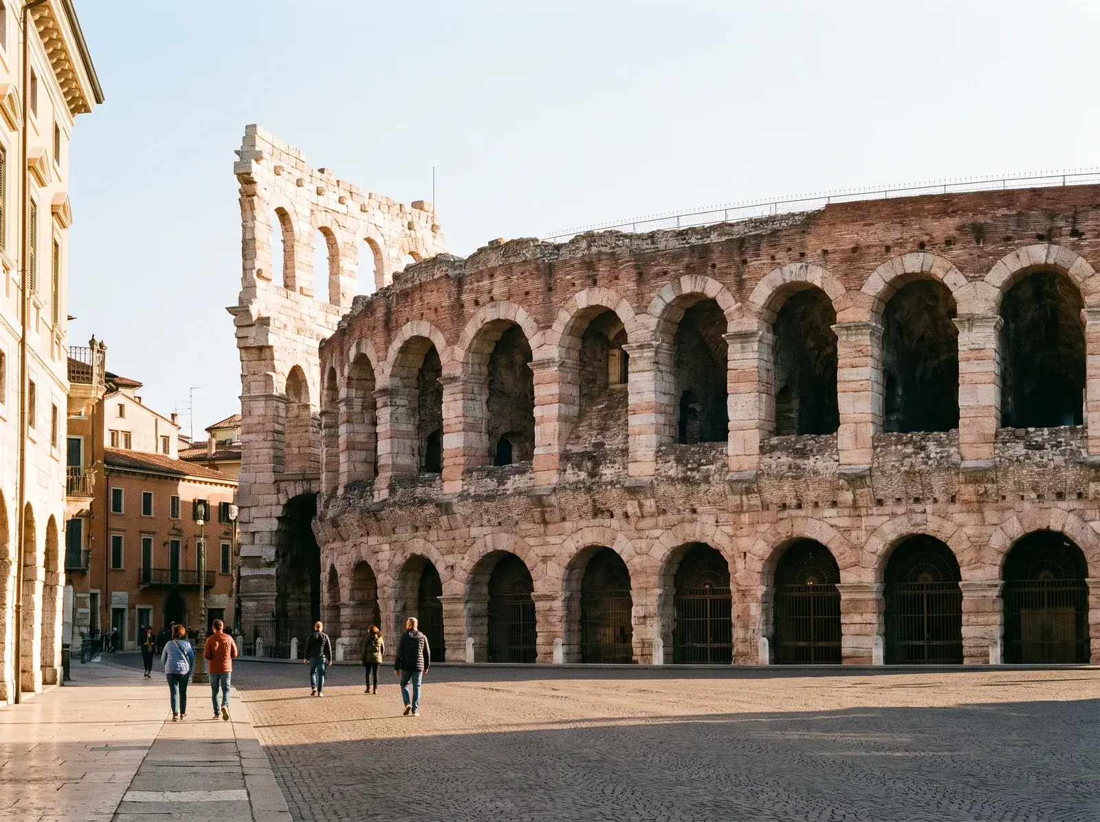 Verona Arena'nın üst üste sıralanan taş kemerleri, önündeki geniş döşeli meydanda yürüyen insanların arkasında uzanıyor.