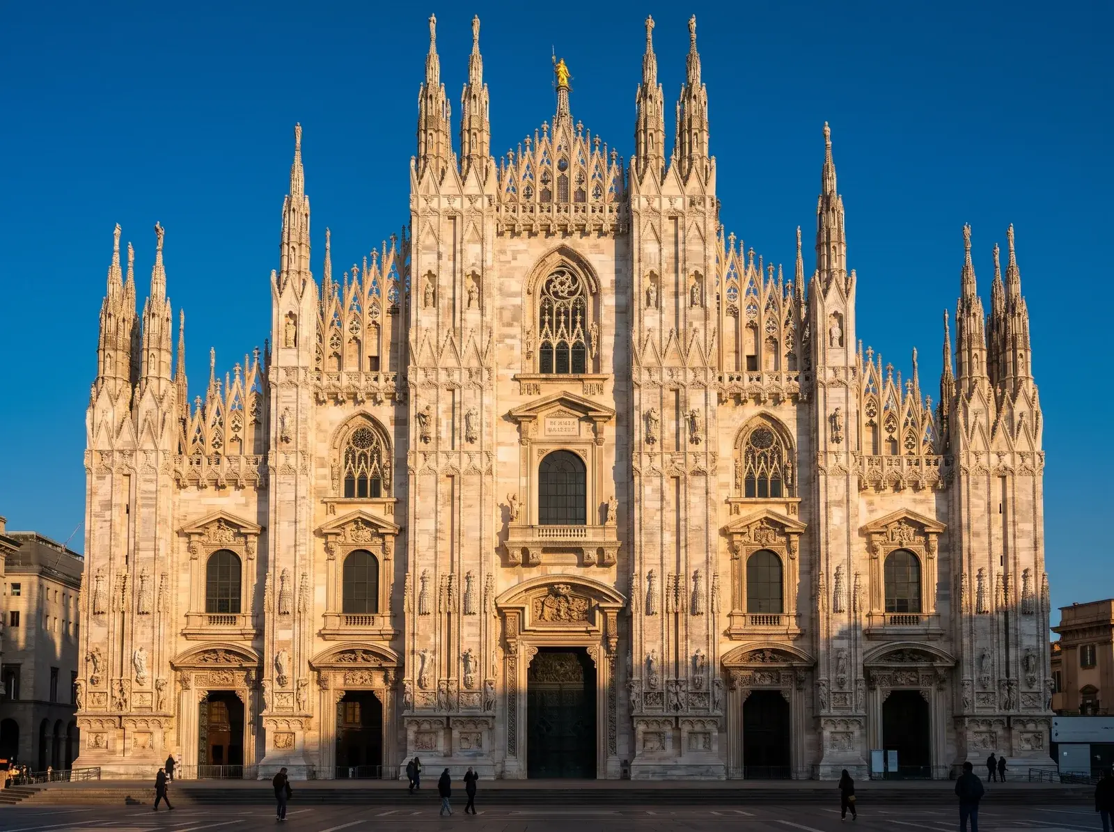Milano Duomo'nun sivri kuleli ve işlemeli cephesi güneşle aydınlanırken önündeki gölgeli meydanda insanlar yürüyor.
