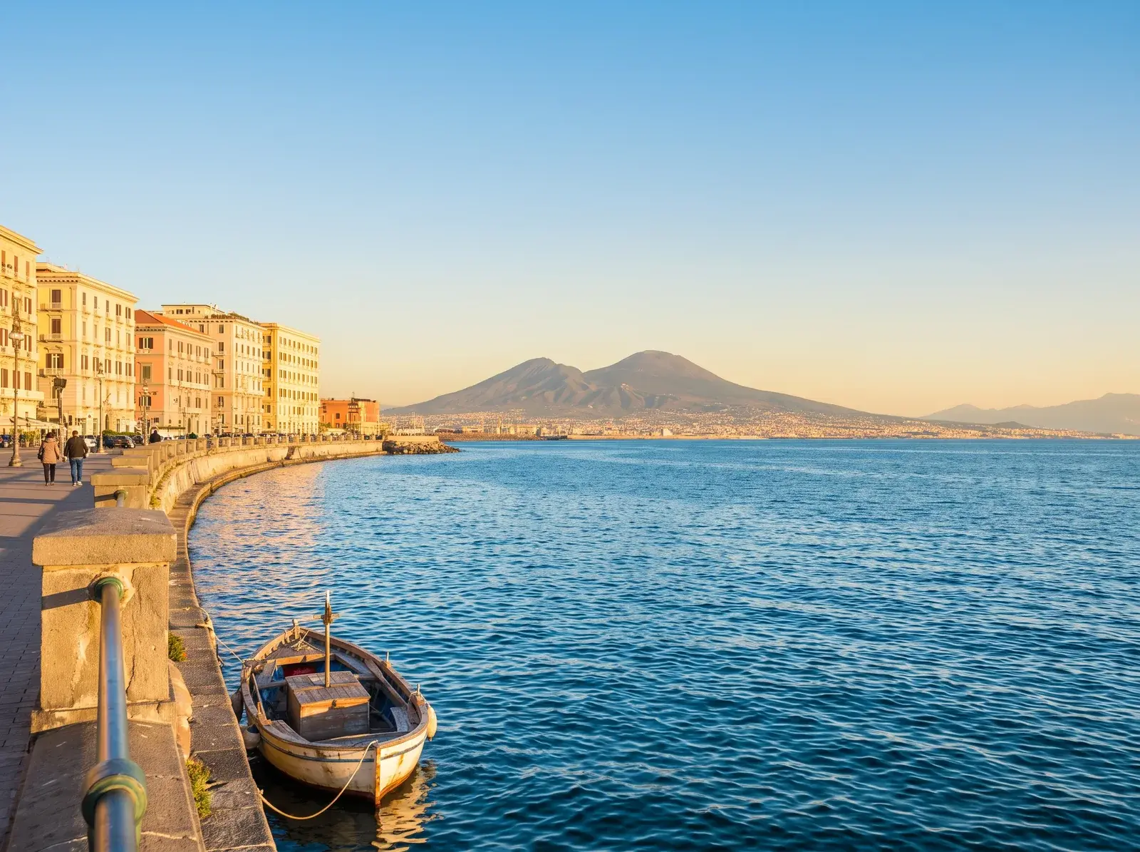 Napoli kıyısında binalar ve bağlı bir kayık, mavi denizin ardında ise Vezüv'ün silueti görülüyor.
