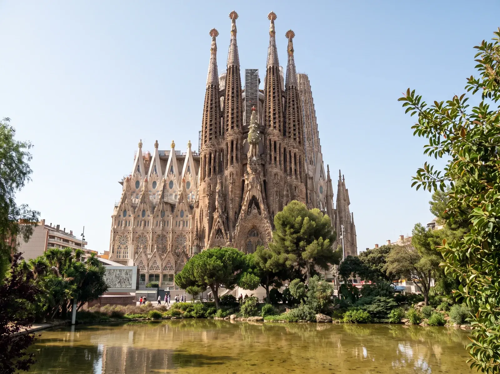 Sagrada Familia'nın işlenmiş taş cephesi ve kuleleri, ağaçlar arasındaki gölete yansıyor.